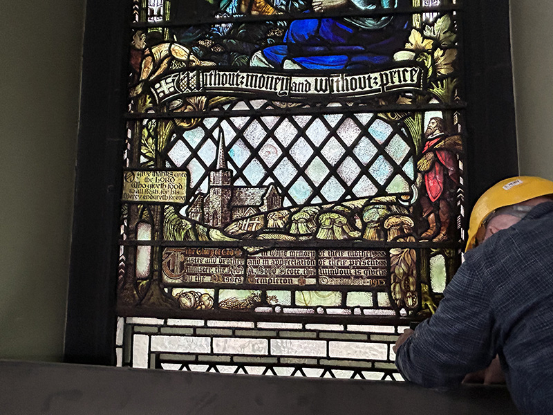 A person wearing a hard-hat working on a historic stained glass window with people, a church and many other words and motifs on it.