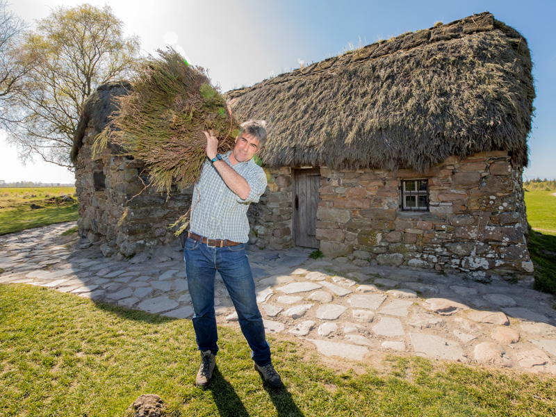 A person wearing a shirt and jeans smiling, while holding a large bundle of thatching material on their shoulder. Behind them is a thatched, stone cottage with a wooden door.