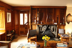A living room with lots of wooden features including wall panelling, a door, tables and chairs and a large wooden fireplace surround. The fireplace itself is a small, triangular, iron fire with a stone surround.