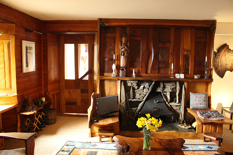 A living room with lots of wooden features including wall panelling, a door, tables and chairs and a large wooden fireplace surround. The fireplace itself is a small, triangular, iron fire with a stone surround.