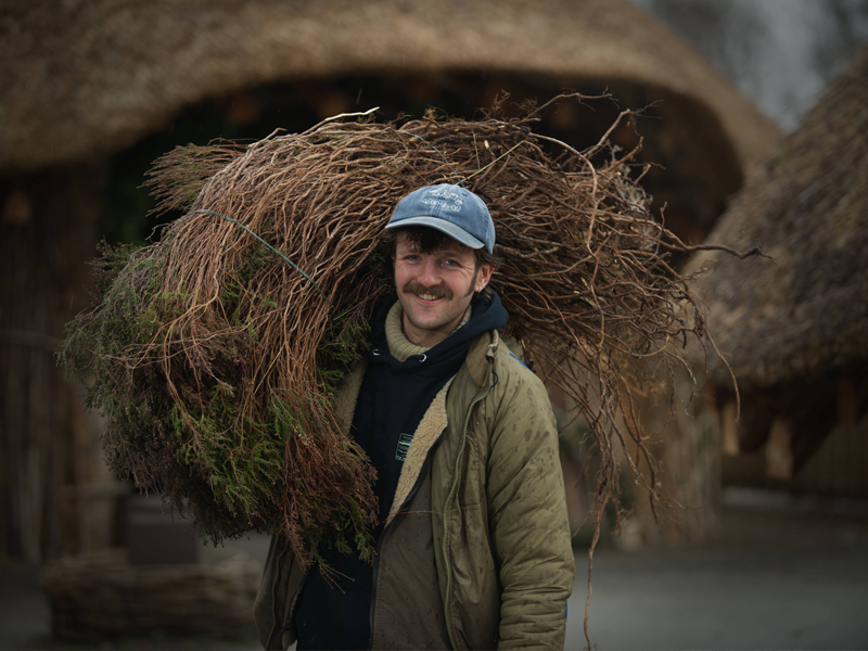 A person with a cap and jacket, standing outside, holding a large pile of thatching material over their shoulder