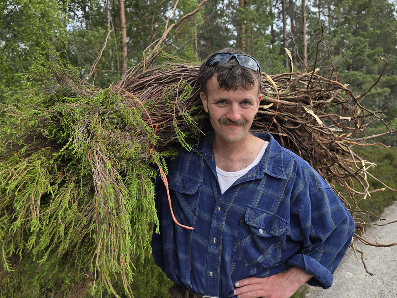 A person wearing sunglasses and a checked shirt smiling while holding a large bunch of heather over their shoulder