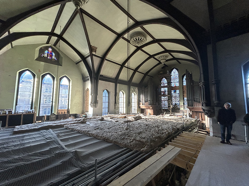 A historic church with a tall, circular ceiling and long stained glass windows. There are chains suspended from the ceiling and large sheets of material on the floor.