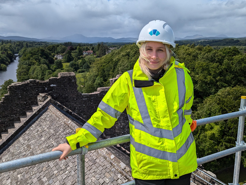 A person with blonde hair, wearing a hard hat and high vis yellow jacket, standing on top of scaffolding
