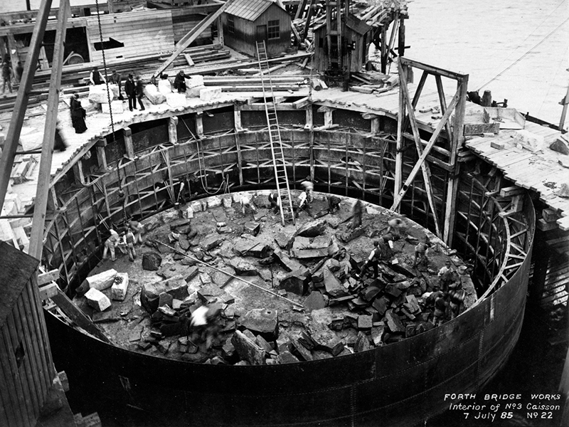 The construction of a large, round structure, with a tall st of ladders and building materials inside the structure. Workers are standing outside it with materials.