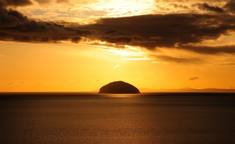 A large mound protruding from the sea. The whole sky is orange as the sun sets.