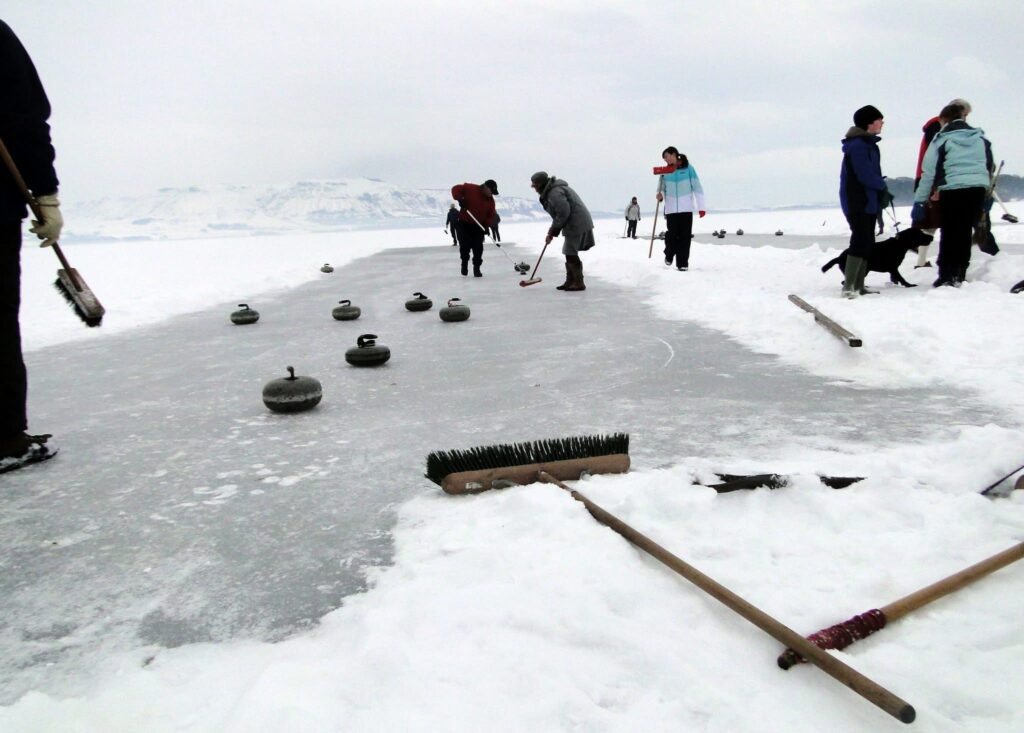 A group of people curling with brushes on a frozen loch, with snowy hills in the background.
