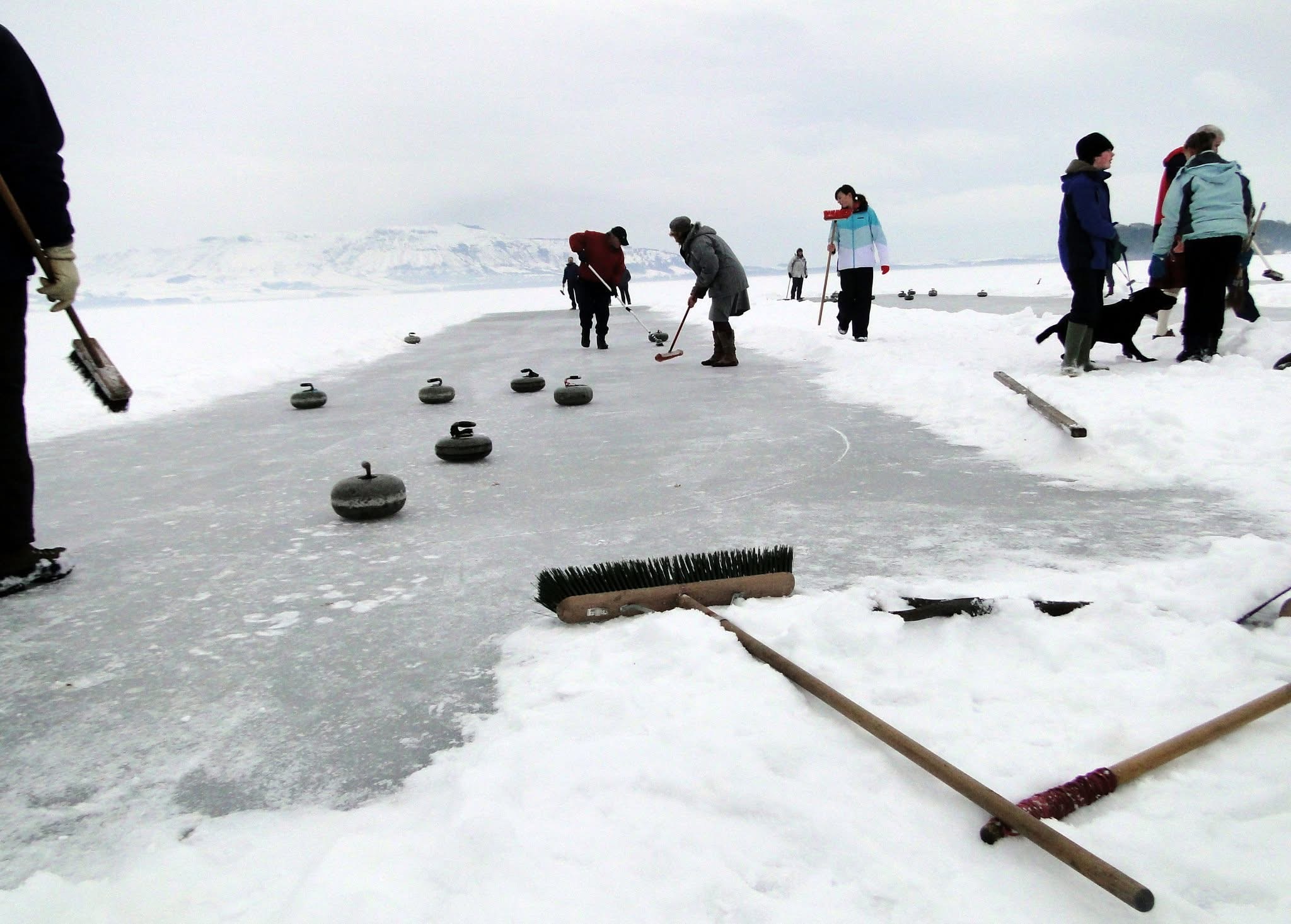 A group of people curling with brushes on a frozen loch, with snowy hills in the background.