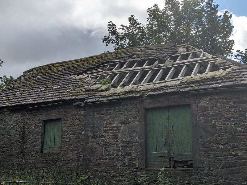 A stone building with a large section of broken roof which has missing flagstones exposing the roof timbers