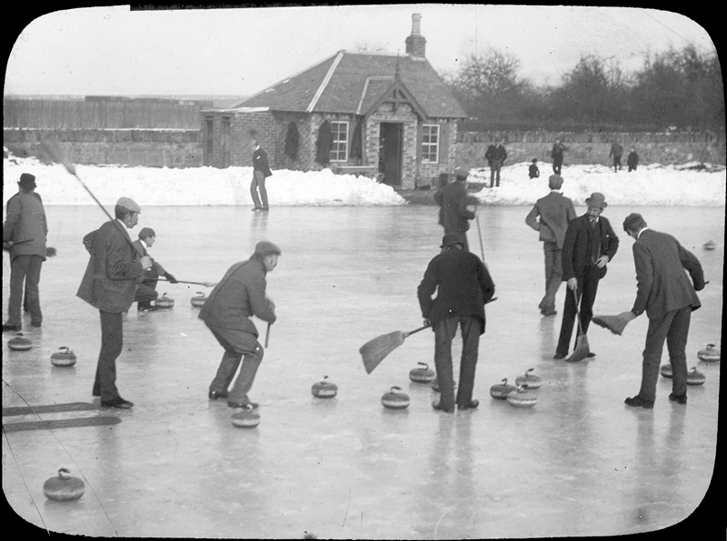 A group of men in suits and hats, curling on an outdoor rink using brooms