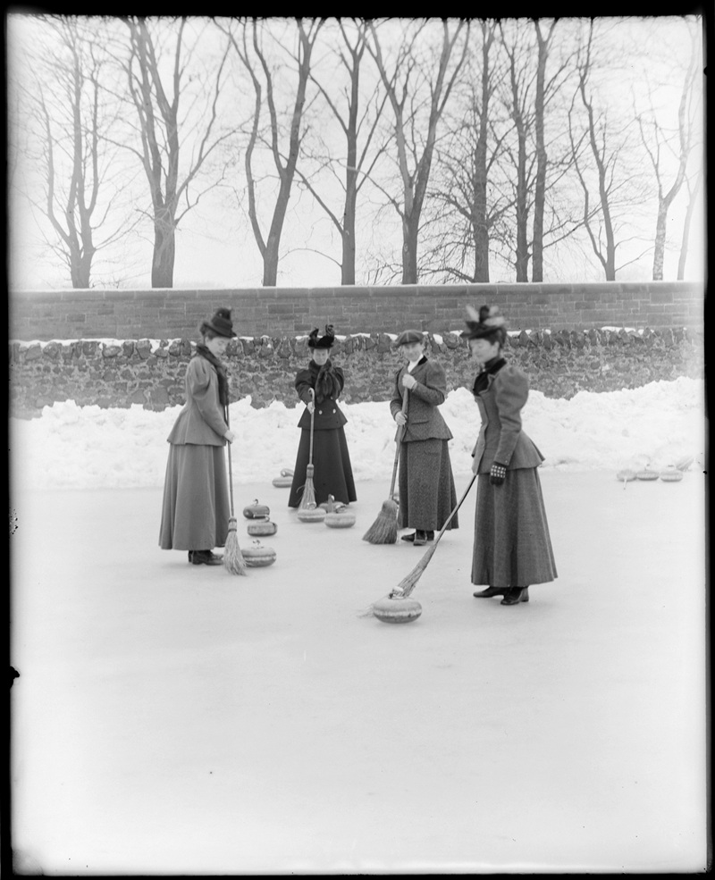 A group of women wearing long skirts, hats and blazers, curling with brooms on an outdoor rink