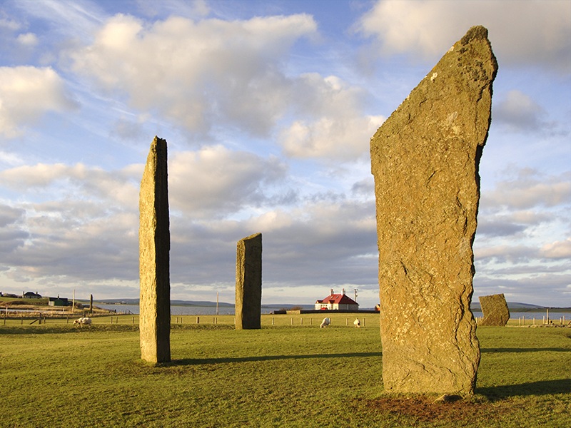 Three tall standing stones in a field and one smaller one, by the coast with a house in the background