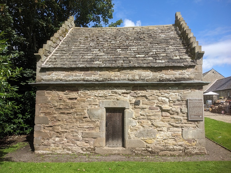 A rectangular, blonde sandstone building with a crow-stepped gable and flagstone roof. A large wooden door serves as an entrance.