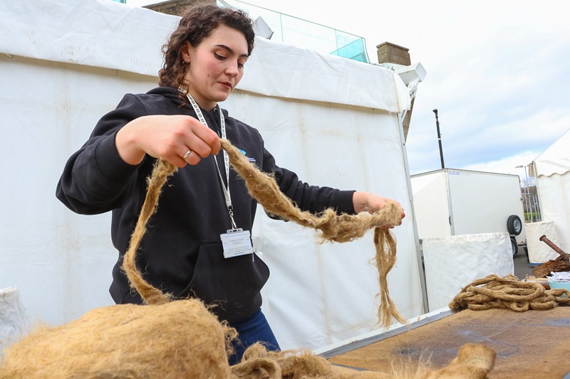 A person wearing a black Historic Environment Scotland hoody, holding up a long piece of oakum at a table in a workshop setting.