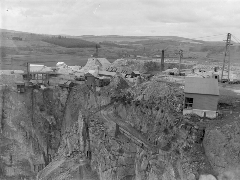 A quarry, with various small wooden buildings and telegraph poles at the top of the rock.