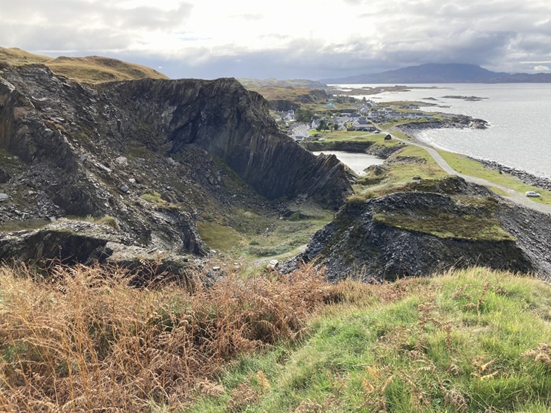 A quarry that is no longer in use, some of it is covered with grass and part of it is filled in with water. There is a small village in the background.