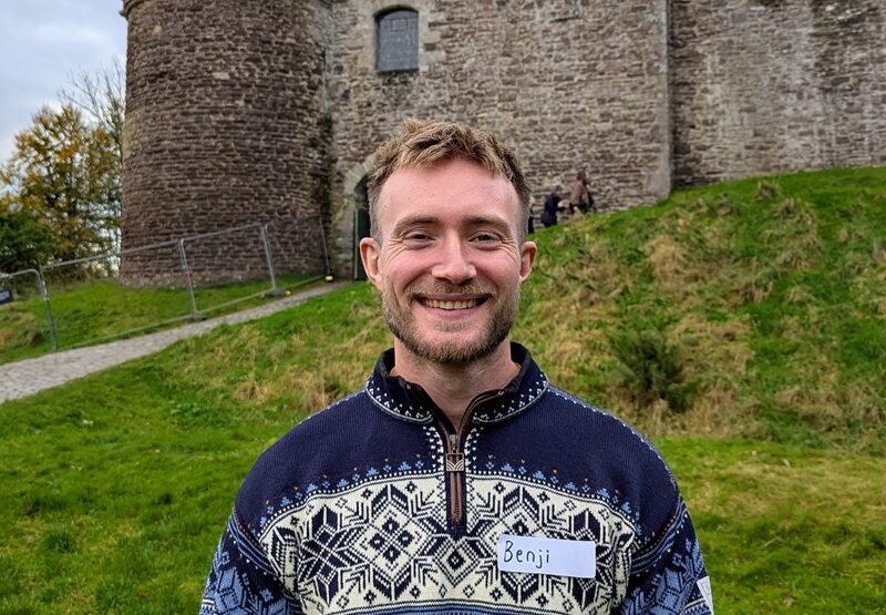 A person wearing a nordic-pattern jumper, smiling widely, in front of Doune Castle