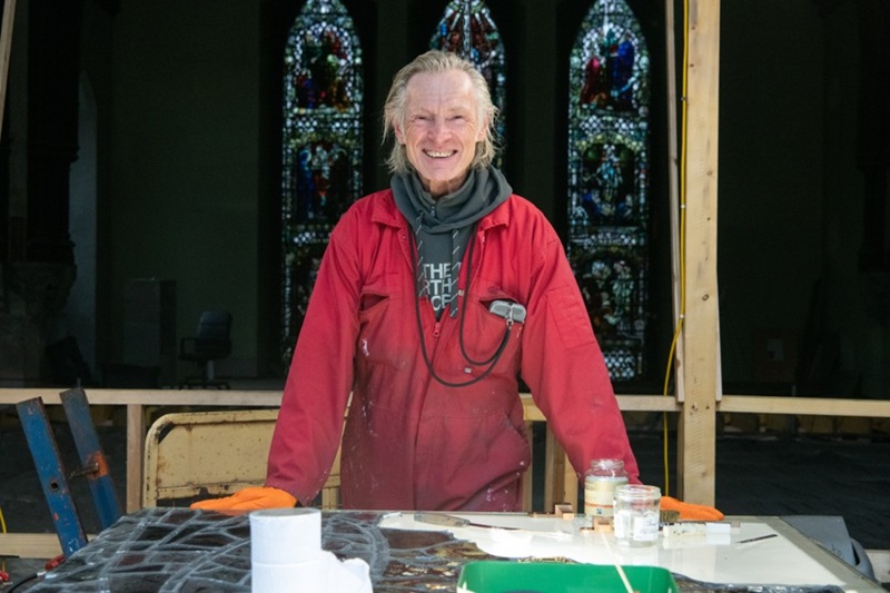 A man, smiling, standing in a historic church in front of large stained glass windows. There is a table in front of him, with a stained glass window work-in-progress on the table