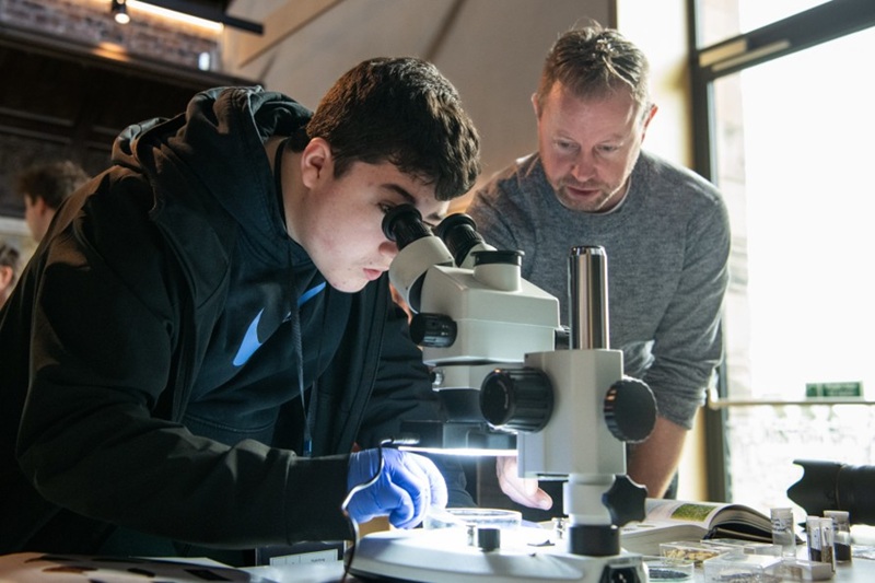 A school pupil looking into a microscope, while an adult talks them through what they're seeing.