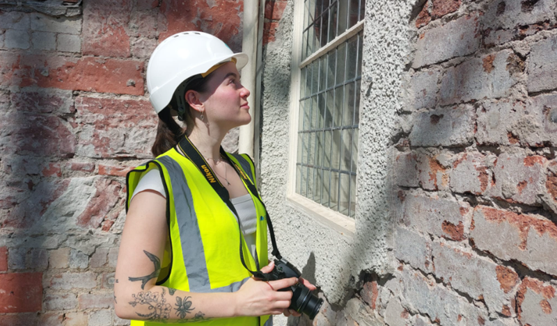 A person wearing a high vis vest and a hard hat looking at a brick wall with damaged harling, holding a camera.