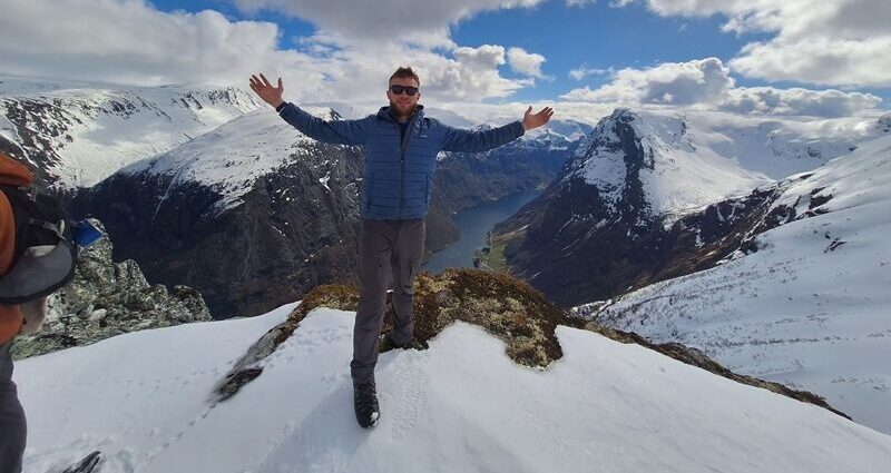 A person wearing a down jacket and sunglasses, holding their arms up on top of a snowy mountain, with a river in the distance below.