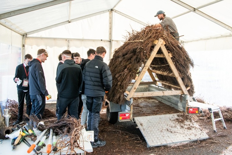 A group of school pupils in a gazebo, listening to a man who is building a thatched roof onto a frame.