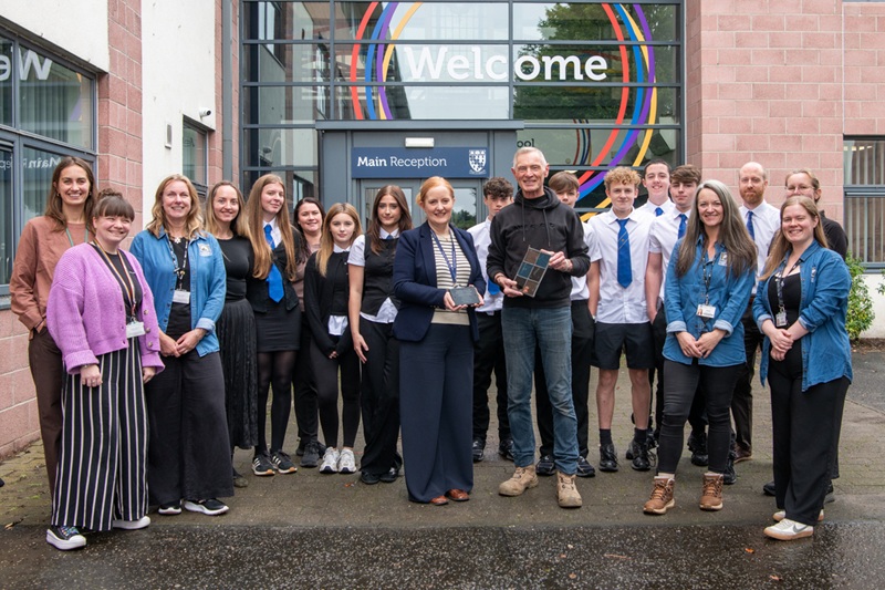 A group of people standing outside a school, including school pupils, holding a stained glass panel and a plaque