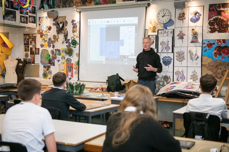 A person standing in front of a classroom, talking to a group of school students who are seated at their desks
