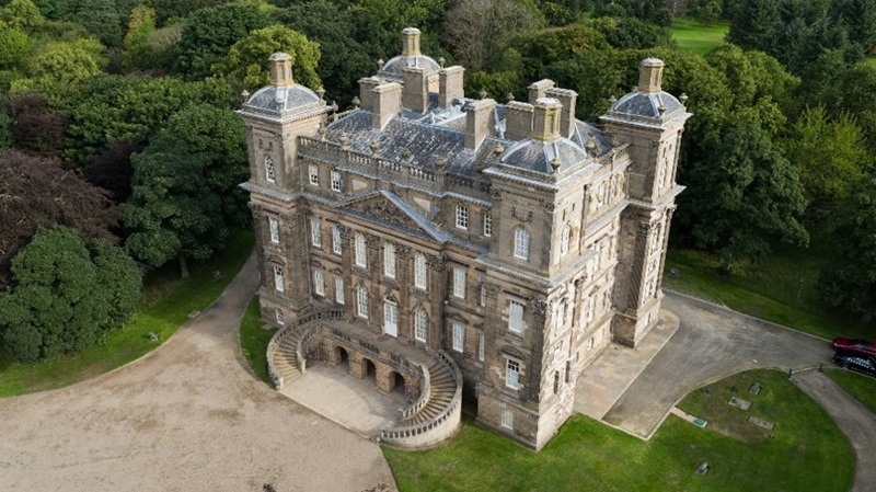 Aerial photograph of a large historic mansion surrounded by dense green trees and a dirt driveway. The mansion features symmetrical architecture with multiple chimneys, a curved front staircase, and a slate roof with dormer windows.